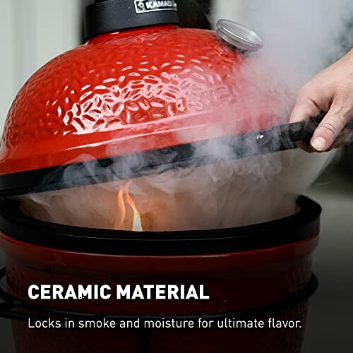 Red ceramic grill with smoke, hand lifting lid.