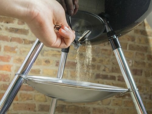 Person cleaning charcoal grill with ash falling into tray.