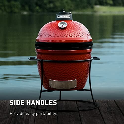 Red Kamado grill with side handles by a lake.